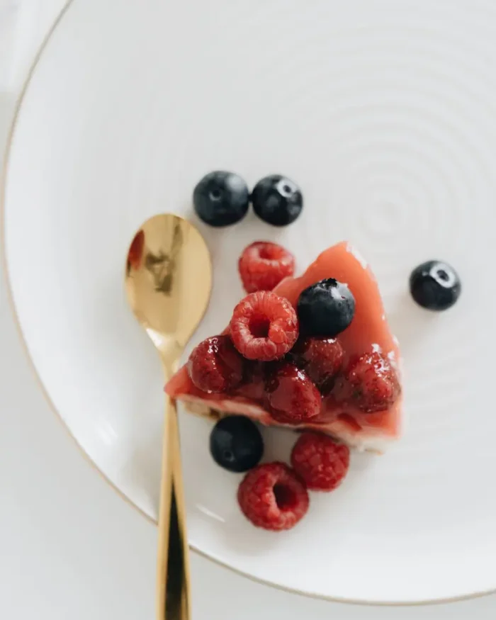 Overhead view of a white plate with a fine gold rim holding a berry cheesecake slice and a gold spoon