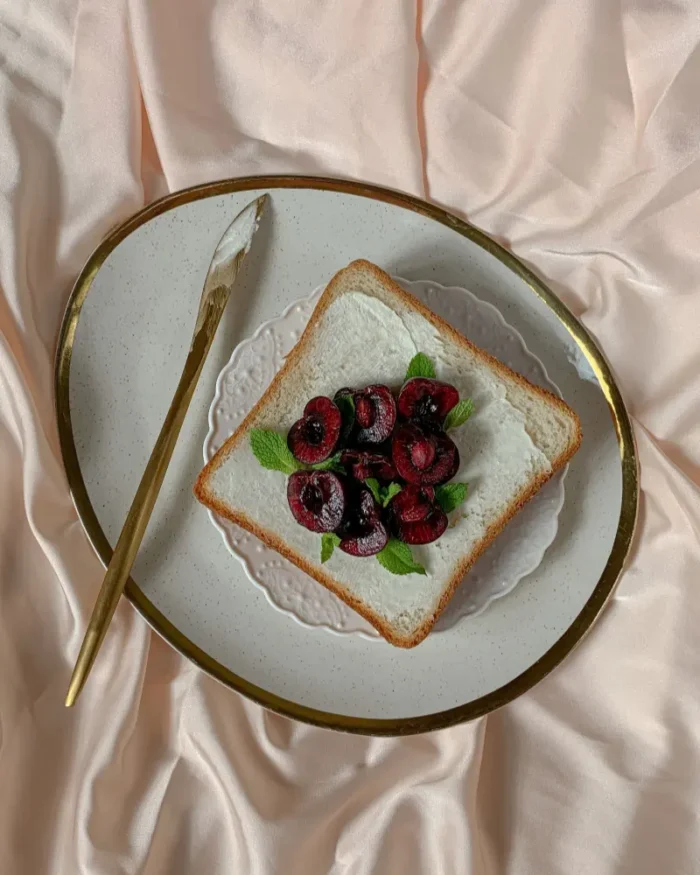 White plate with a gold rim serving toast topped with cherries, styled on blush silk with a gold knife