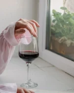 Hand resting on a Lumière crystal wine glass of red wine on a sunny windowsill beside an open book
